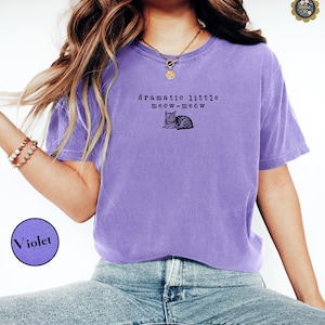May include: A violet short-sleeved t-shirt featuring a black graphic of a cat and the text "dramatic little meow-meow" printed on the chest. The shirt has a crew neckline and a relaxed fit, paired with blue denim jeans.