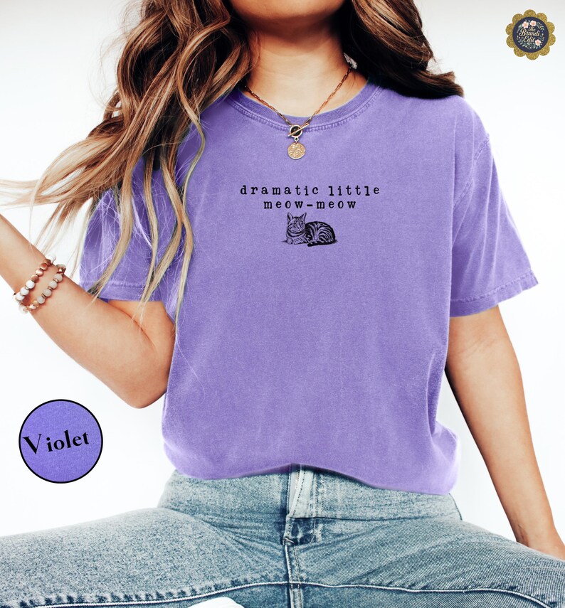 May include: A violet short-sleeved t-shirt featuring a black graphic of a cat and the text "dramatic little meow-meow" printed on the chest. The shirt has a crew neckline and a relaxed fit, paired with blue denim jeans.