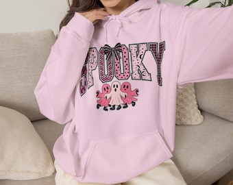 Może przedstawiać: Jasnoróżowa bluza z kapturem z nadrukiem graficznym z przodu. Napis SPOOKY wykonany jest dużymi literami z różnymi różowymi i czarnymi wzorami. Poniżej napisu znajdują się trzy rysunkowe duszki w odcieniach różu i bieli.