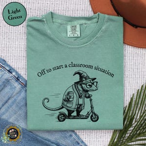 May include: A light green t-shirt featuring a black graphic of an opossum wearing a witch hat, glasses, and a cardigan while riding a scooter and holding a coffee cup. The text above reads, Off to start a classroom situation.