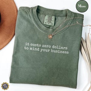 May include: A moss green t-shirt with the text "it costs zero dollars to mind your business" in white. A gold necklace with a pendant rests on the shirt. The shirt is labeled "Comfort Colors" and is size small.
