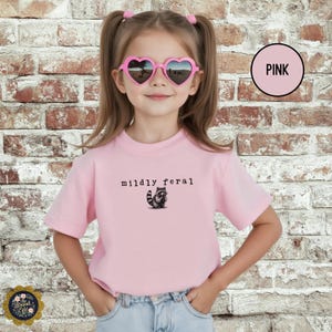 Puede incluir: Una camiseta de manga corta de color rosa claro con un peque&ntilde;o gr&aacute;fico de un mapache y el texto "mildly feral" impreso en negro. La camiseta se combina con unos vaqueros azul claro y unas gafas de sol rosas en forma de coraz&oacute;n.