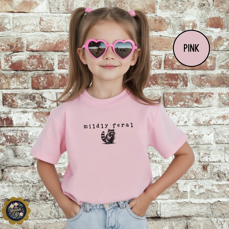 Puede incluir: Una camiseta de manga corta de color rosa claro con un peque&ntilde;o gr&aacute;fico de un mapache y el texto "mildly feral" impreso en negro. La camiseta se combina con unos vaqueros azul claro y unas gafas de sol rosas en forma de coraz&oacute;n.