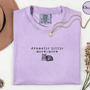 May include: A light purple t-shirt featuring the black embroidered text "dramatic little meow-meow" above a small illustration of a cat. The shirt has a crew neckline and a Comfort Colors brand label on the inside collar, size XL.
