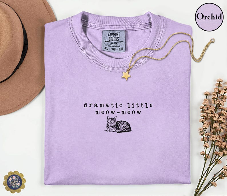 May include: A light purple t-shirt featuring the black embroidered text "dramatic little meow-meow" above a small illustration of a cat. The shirt has a crew neckline and a Comfort Colors brand label on the inside collar, size XL.