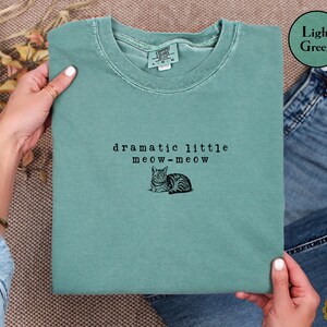 May include: A light green crewneck t-shirt featuring black embroidered text that reads dramatic little meow-meow above a small black embroidered illustration of a cat lying down. The shirt has a ribbed collar and a tag indicating size large.