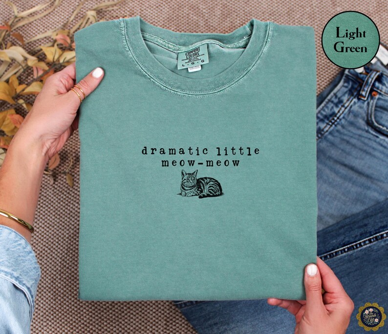 May include: A light green crewneck t-shirt featuring black embroidered text that reads dramatic little meow-meow above a small black embroidered illustration of a cat lying down. The shirt has a ribbed collar and a tag indicating size large.