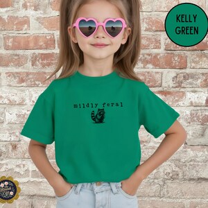Puede incluir: Una camiseta de manga corta color verde Kelly con un gr&aacute;fico negro de un peque&ntilde;o mapache y el texto "mildly feral" impreso en una fuente estilo m&aacute;quina de escribir en el pecho. Se lleva con vaqueros azul claro y gafas de sol en forma de coraz&oacute;n.