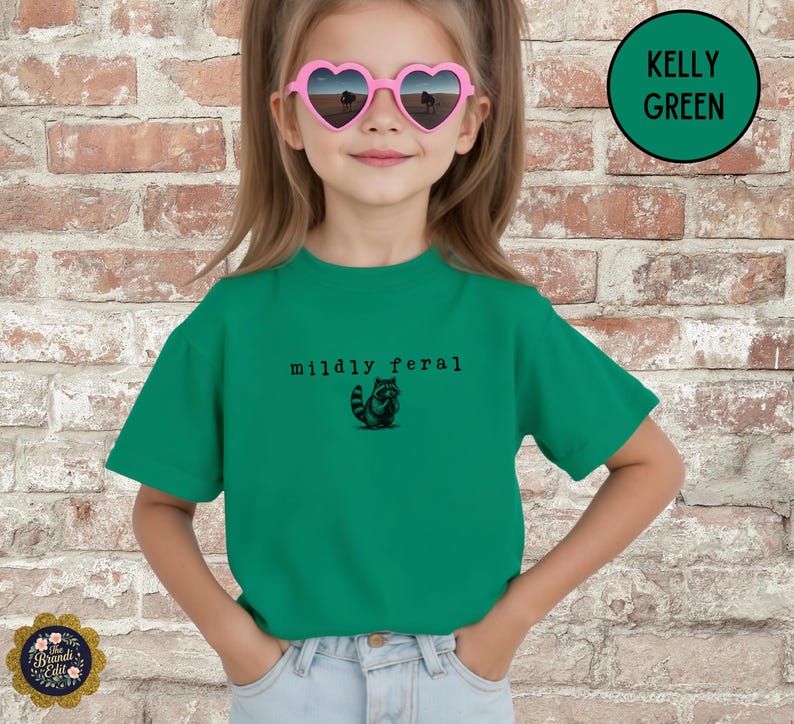 Puede incluir: Una camiseta de manga corta color verde Kelly con un gr&aacute;fico negro de un peque&ntilde;o mapache y el texto "mildly feral" impreso en una fuente estilo m&aacute;quina de escribir en el pecho. Se lleva con vaqueros azul claro y gafas de sol en forma de coraz&oacute;n.