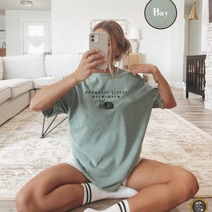 May include: A sage green short-sleeved t-shirt featuring a black graphic of a cat and the text "dramatic little meow-meow" printed on the chest. The shirt has a crew neckline and a relaxed fit, with a small circular colour swatch labelled "Bay".