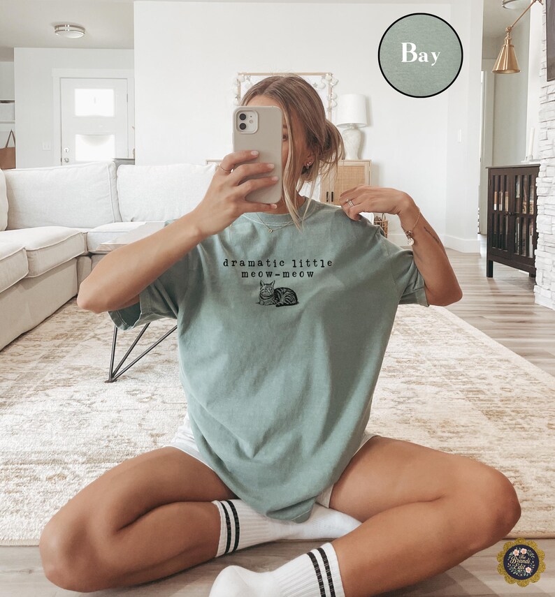 May include: A sage green short-sleeved t-shirt featuring a black graphic of a cat and the text "dramatic little meow-meow" printed on the chest. The shirt has a crew neckline and a relaxed fit, with a small circular colour swatch labelled "Bay".