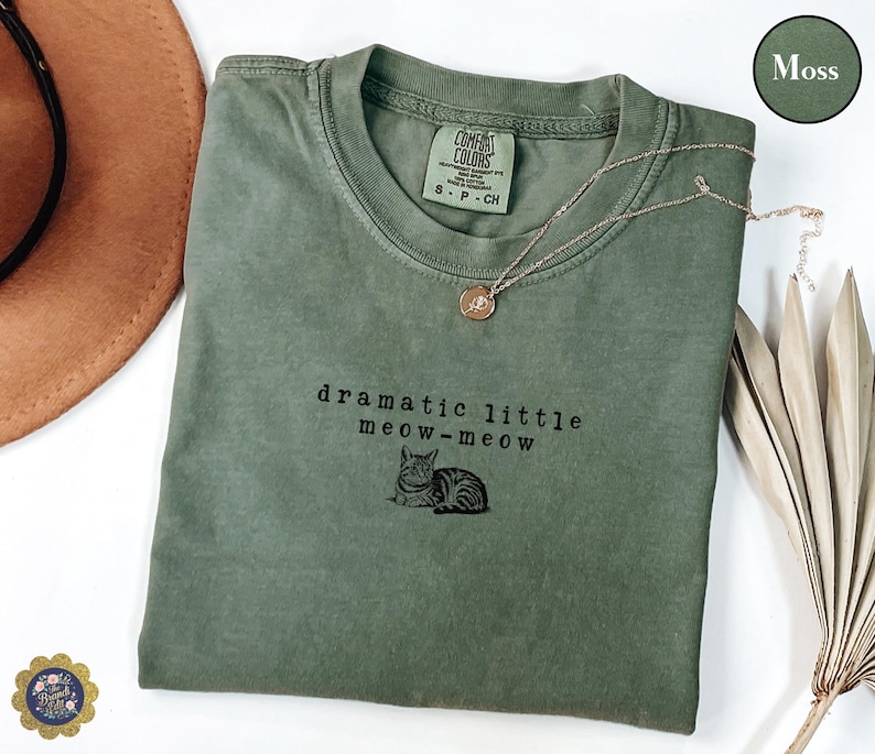 May include: A moss green t-shirt with the text "dramatic little meow-meow" and a cat illustration. A gold necklace with a pendant is draped on the shirt. The shirt is folded and displayed on a white surface.