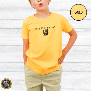 Puede incluir: Una camiseta de manga corta de color oro con un peque&ntilde;o gr&aacute;fico negro de un mapache y el texto "mildly feral" impreso en una fuente estilo m&aacute;quina de escribir en el centro del pecho. La camiseta tiene cuello redondo.