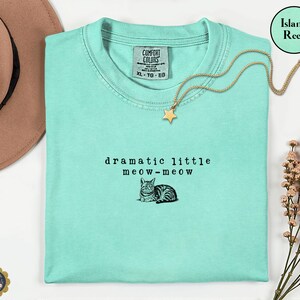 May include: An island reef coloured t-shirt featuring the black text "dramatic little meow-meow" and a small illustration of a cat. The shirt has a Comfort Colors brand label at the neckline, indicating a size XL, and is made of 100% cotton fabric.