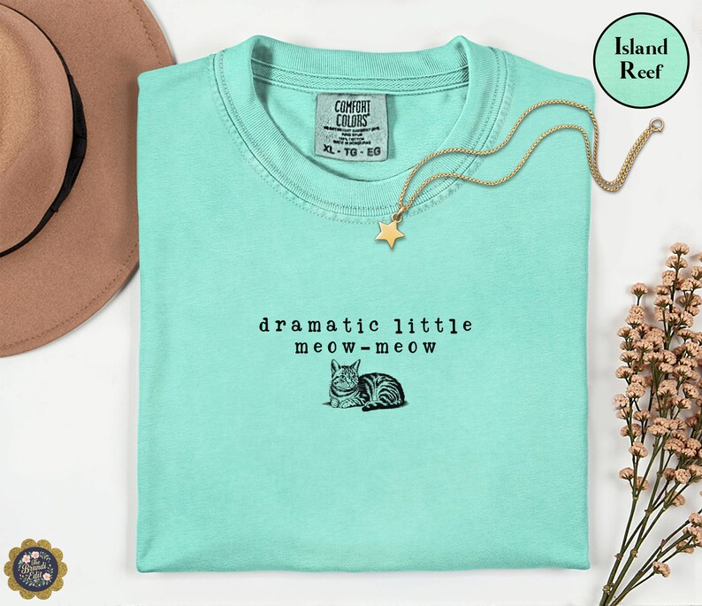 May include: An island reef coloured t-shirt featuring the black text "dramatic little meow-meow" and a small illustration of a cat. The shirt has a Comfort Colors brand label at the neckline, indicating a size XL, and is made of 100% cotton fabric.