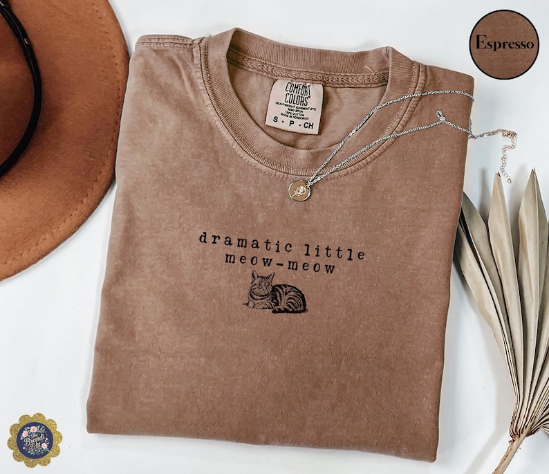 May include: An espresso-coloured t-shirt featuring the embroidered text "dramatic little meow-meow" above a small illustration of a tabby cat. The shirt has a crew neckline and a Comfort Colors brand label inside the collar.