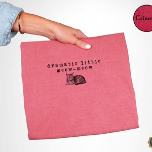 May include: A crimson t-shirt featuring the black text "dramatic little meow-meow" above a black and white illustration of a cat. The fabric has a heathered texture, and the design is centred on the front of the garment.