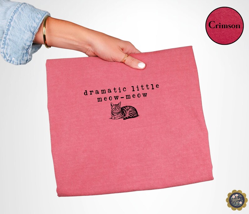 May include: A crimson t-shirt featuring the black text "dramatic little meow-meow" above a black and white illustration of a cat. The fabric has a heathered texture, and the design is centred on the front of the garment.
