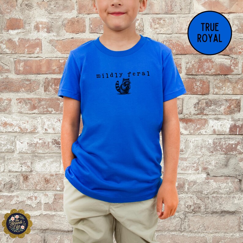 Puede incluir: Una camiseta de manga corta en color azul real brillante que presenta un peque&ntilde;o gr&aacute;fico negro de un mapache y el texto "mildly feral" impreso en una fuente negra sencilla en el centro del pecho. Se combina con pantalones de color beige claro.