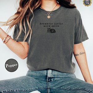 May include: A charcoal grey short-sleeve t-shirt featuring a black graphic of a cat and the text "dramatic little meow-meow" printed on the chest. The shirt has a crew neckline and a relaxed fit, shown in the colour labelled as Pepper.