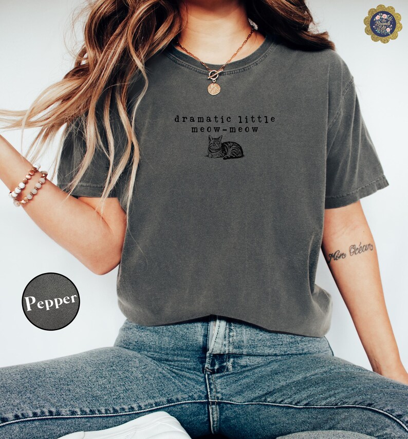 May include: A charcoal grey short-sleeve t-shirt featuring a black graphic of a cat and the text "dramatic little meow-meow" printed on the chest. The shirt has a crew neckline and a relaxed fit, shown in the colour labelled as Pepper.