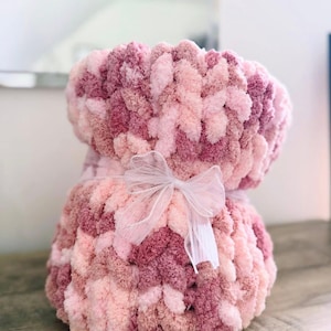 Puede incluir: Una manta suave tejida a mano en tonos rosa y malva, atada con una delicada cinta blanca. La manta está hecha de hilo grueso y texturizado, creando una apariencia acogedora e invitadora. Ideal para un baby shower o un regalo.