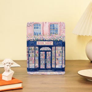Puede incluir: Una impresión rectangular decorativa de una pintoresca librería. La tienda tiene una fachada rosa, adornos azules y un letrero que dice "BOOK SHOP". Las ventanas están llenas de libros y la parte superior está adornada con flores. Un pequeño busto y un libro están en primer plano.