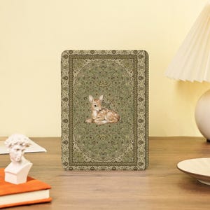 Könnte beinhalten: Ein rechteckiger Dekorationsartikel mit einer detaillierten Illustration eines Rehs, das auf einem grün-beigen Teppich mit Muster ruht. Das Reh ist braun mit weißen Flecken. Der Teppich hat ein Blumenmuster.