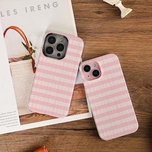 May include: Two pink and white striped phone cases. The cases are on a wooden surface. The cases have a vertical striped pattern. The phone cases are next to a magazine with the text "LES IRENG".