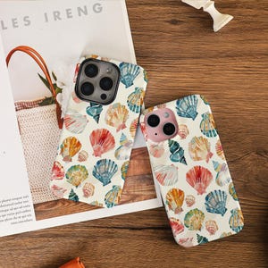May include: Two phone cases featuring a cream backdrop adorned with a pattern of vibrant seashells in red, orange, blue, and green. The cases rest on a white surface, with the text "LES IRENG" visible.