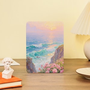 Könnte beinhalten: Ein rechteckiger Kunstdruck zeigt eine Küstenlandschaft mit türkisfarbenen Wellen, die gegen Felsen schlagen. Rosa Blumen blühen an den Klippen, und der Himmel geht von Rosa zu Gelb über, was auf einen Sonnenuntergang hindeutet. Das Kunstwerk wird auf einer Holzoberfläche präsentiert.