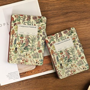 Może przedstawiać: Dwa zeszyty kompozycyjne z kwiatowymi wzorami. Zeszyty mają kremowe tło z wzorem kolorowych kwiatów i zielonych liści. Na każdym zeszycie znajduje się napis "Composition Notebook".