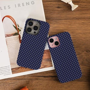 Puede incluir: Dos fundas de teléfono azul marino con lunares blancos. Están sobre una superficie de madera junto a una revista con el texto "LES IRENG". Una tiene un módulo de cámara plateado, la otra rosa.