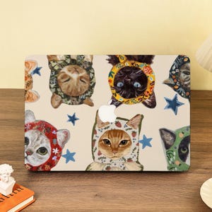 Könnte beinhalten: Eine Laptop-Hülle mit einem skurrilen Design von Katzen mit gemusterten Hüten. Die Katzen sind in verschiedenen Farben und Mustern dargestellt, umgeben von blauen Sternen auf cremefarbenem Hintergrund. Die Hülle hat einen Apple-Logo-Ausschnitt.