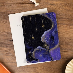 May include: A tablet case with a night sky design. It features a dark blue background, swirling purple and gold patterns, and hanging gold stars. The case rests on a white card with the letters "TO".
