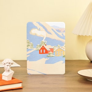 以下が含まれることがあります： 冬の風景イラストが描かれた、角が丸い長方形のケース。赤い家と黄色い家、常緑樹、青い空の下の山々が雪景色の中に描かれています。赤い家の煙突からは煙が上がっています。