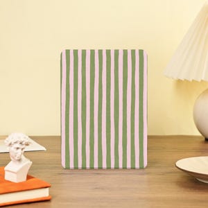 Puede incluir: Un cuaderno rectangular con un patrón de rayas verticales en verde oliva y rosa claro. El cuaderno está de pie sobre una superficie de madera. Un pequeño busto blanco y un libro naranja están en primer plano.
