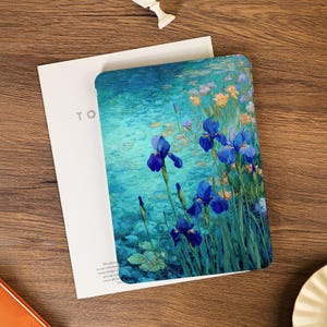 Puede incluir: Una funda para tableta con una pintura de lirios azules en un estanque. La obra de arte tiene un estilo impresionista con tonos de azul, verde y amarillo. La funda está sobre una superficie de madera junto a un libro blanco con las letras "TO".