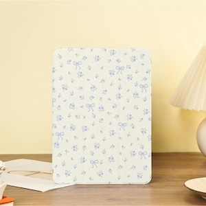 May include: A rectangular, cream-coloured case featuring a floral pattern of small blue flowers and bows. The case is likely for a tablet or similar device, and the design gives it a vintage, feminine aesthetic. The background is a soft yellow.