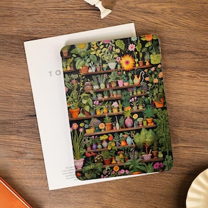May include: A notebook with a detailed illustration of a plant shelf, filled with potted plants and flowers. The notebook is open, with the text "TO" visible on the left page. The cover displays a vibrant array of greens, oranges, and pinks.