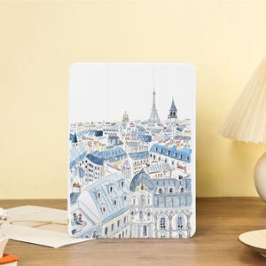 Könnte beinhalten: Weiße Tablet-Hülle mit einer Aquarellillustration einer Stadtlandschaft, einschließlich des Eiffelturms und Gebäude mit blauen Dächern. Das Kunstwerk zeigt eine charmante Szene mit einem Paar auf einem Dach. Die Hülle liegt auf einer Holzoberfläche.