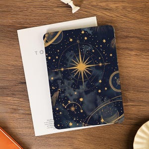 May include: A navy blue notebook with a celestial design featuring gold stars, planets, and a compass rose. The notebook is open, revealing a white interior. The word "TO" is visible on the left side of the notebook.