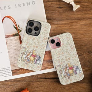 May include: Two phone cases featuring a floral design and a cartoon illustration of Winnie the Pooh and friends. The cases are beige with a repeating pattern of small pink and green flowers.