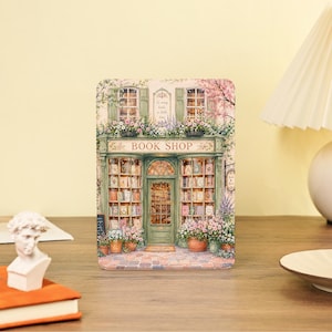 May include: A rectangular metal tin featuring a detailed illustration of a green storefront with the text BOOK SHOP above the entrance. The shop has large windows filled with books, flower boxes, and a cobblestone path in front of the building.