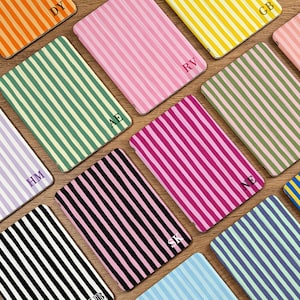 May include: A collection of personalized notebooks with vertical stripes in various colors, including pink, green, yellow, and black. Each notebook features a different initial in the corner. The notebooks are arranged on a wooden surface.