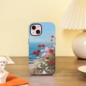 May include: A phone case with a coastal landscape design. The case depicts red poppies, pink and white flowers, and a blue sea under a clear sky. The phone case has a black camera housing.