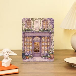 May include: A rectangular decorative sign featuring a detailed illustration of a purple storefront titled The Blooming Bookshop. The facade is adorned with climbing wisteria, potted flowers, and large glass windows displaying shelves filled with books.