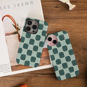 May include: Two mobile phone cases featuring a green and pale green chequered design. They are displayed on a wooden surface, next to a magazine with the text "LES IRENG". One case has a black camera, the other a pink camera.