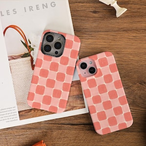 May include: Two phone cases with a pink and peach checkerboard pattern. The cases are on a wooden surface, with a magazine and a bag in the background. The cases have camera cutouts.