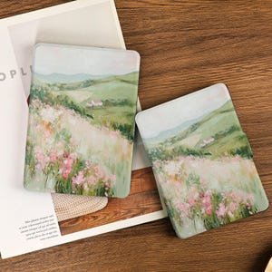 以下が含まれることがあります： 風景画が描かれた長方形のケースが2つ。 ピンクの花畑、緑の丘、薄い青い空の下の小さな白い家を描いたアートワークです。 ケースはおそらくタブレットまたは電子書籍リーダー用です。